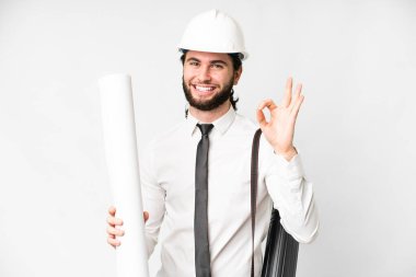 Young architect man with helmet and holding blueprints over isolated white background showing ok sign with fingers
