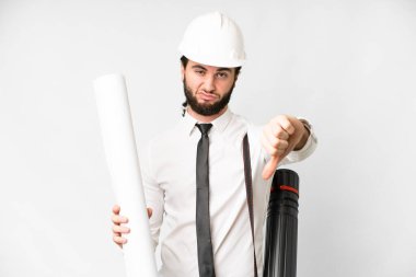Young architect man with helmet and holding blueprints over isolated white background showing thumb down with negative expression