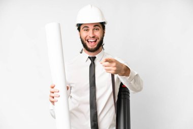 Young architect man with helmet and holding blueprints over isolated white background surprised and pointing front