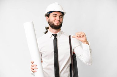 Young architect man with helmet and holding blueprints over isolated white background proud and self-satisfied