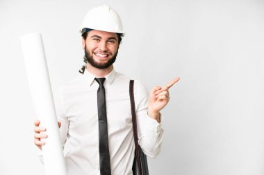 Young architect man with helmet and holding blueprints over isolated white background pointing to the side to present a product
