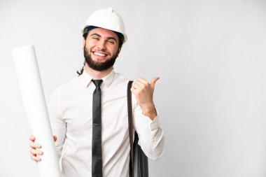 Young architect man with helmet and holding blueprints over isolated white background pointing to the side to present a product