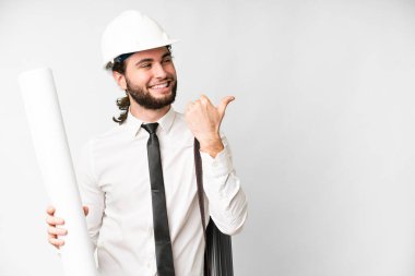 Young architect man with helmet and holding blueprints over isolated white background pointing to the side to present a product