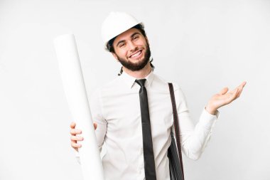 Young architect man with helmet and holding blueprints over isolated white background extending hands to the side for inviting to come