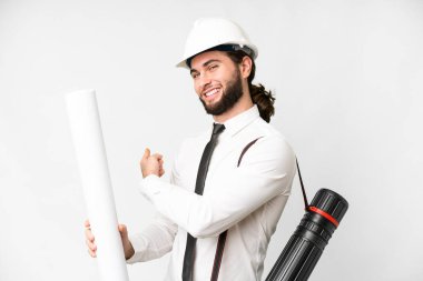 Young architect man with helmet and holding blueprints over isolated white background pointing back