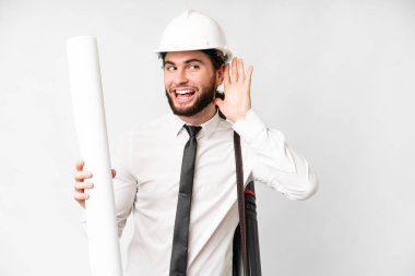 Young architect man with helmet and holding blueprints over isolated white background listening to something by putting hand on the ear