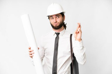 Young architect man with helmet and holding blueprints over isolated white background with fingers crossing and wishing the best