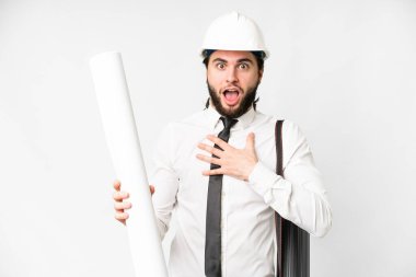 Young architect man with helmet and holding blueprints over isolated white background surprised and shocked while looking right