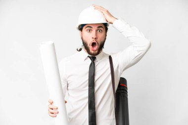 Young architect man with helmet and holding blueprints over isolated white background doing surprise gesture while looking to the side