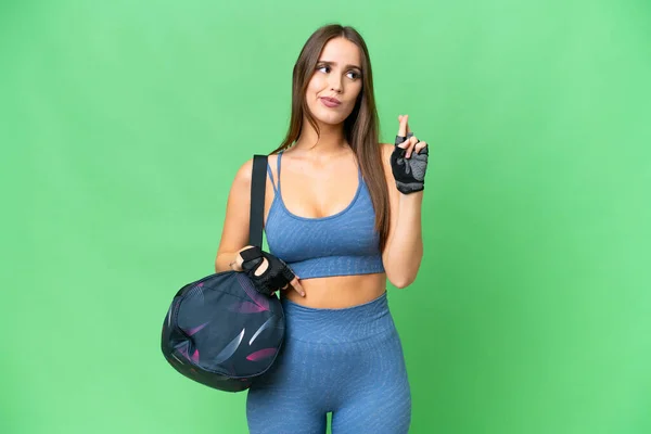 Young sport woman with sport bag over isolated chroma key background with fingers crossing and wishing the best