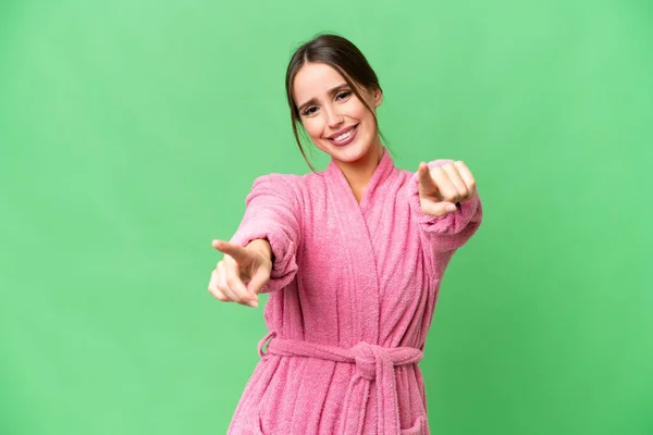 Young beautiful woman in a bathrobe over isolated chroma key background points finger at you while smiling