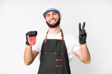 Butcher man wearing an apron and serving fresh cut meat over isolated white background smiling and showing victory sign