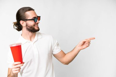 Young handsome man holding soda while watching a 3D movie over isolated white background pointing to the side to present a product
