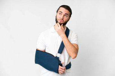 Young handsome man with broken arm and wearing a sling over isolated white background having doubts