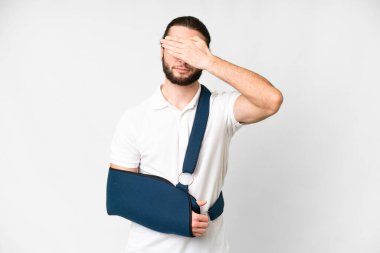 Young handsome man with broken arm and wearing a sling over isolated white background covering eyes by hands. Do not want to see something