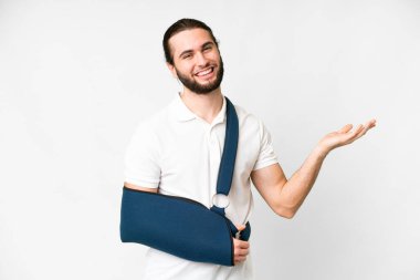 Young handsome man with broken arm and wearing a sling over isolated white background extending hands to the side for inviting to come