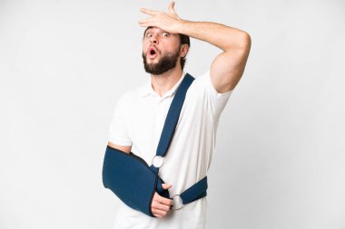 Young handsome man with broken arm and wearing a sling over isolated white background doing surprise gesture while looking to the side