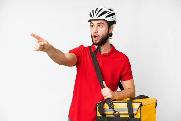 Young delivery man with thermal backpack over isolated white background pointing away