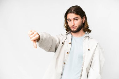 Soyutlanmış beyaz arka planda genç yakışıklı bir adam negatif ifadeyle baş parmağını aşağı gösteriyor.