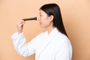 Bej arka planda izole edilmiş genç Çinli kadın elinde makyaj fırçası tutuyor.