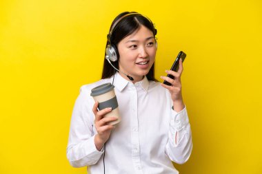 Tele pazarlamacı Çinli bir kadın sarı arka planda izole edilmiş bir kulaklıkla çalışıyor. Elinde kahve ve cep telefonu var.