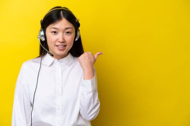 Sarı arka planda izole edilmiş kulaklıkla çalışan pazarlamacı Çinli kadın bir ürünü sunmak için yan tarafı işaret ediyor.