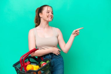 Young woman holding a shopping basket full of food isolated on green background pointing finger to the side and presenting a product