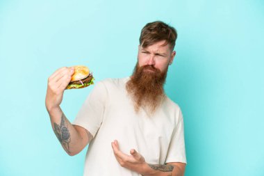 Kızıl saçlı, uzun sakallı bir adam elinde burgerle mavi arka planda üzgün bir ifadeyle izole edilmiş.