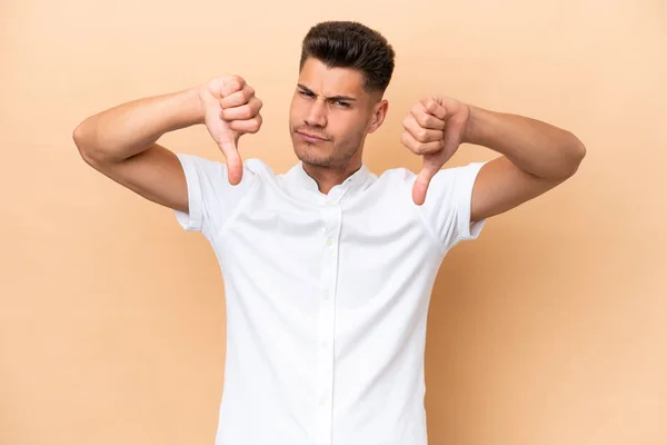 Young caucasian man isolated on beige background showing thumb down with two hands