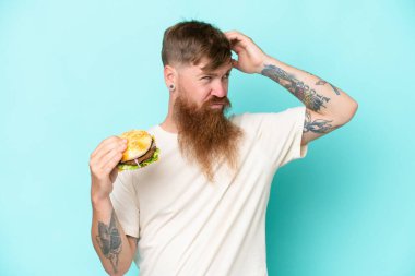 Redhead man with long beard holding a burger isolated on blue background having doubts and with confuse face expression