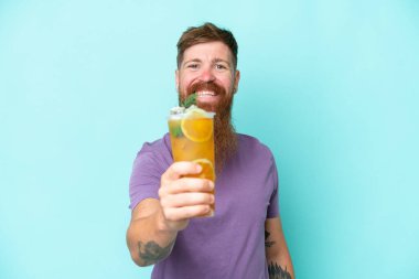 Redhead man with long beard holding a cocktail isolated on blue background with happy expression