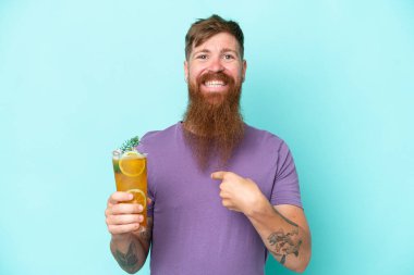 Redhead man with long beard holding a cocktail isolated on blue background with surprise facial expression