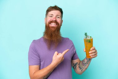 Redhead man with long beard holding a cocktail isolated on blue background and pointing it