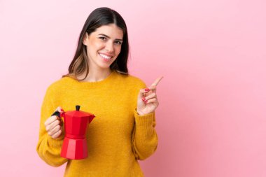 Pembe arka planda izole edilmiş kahve makinesini tutan genç İtalyan kadın bir ürünü sunmak için kenarı işaret ediyor.