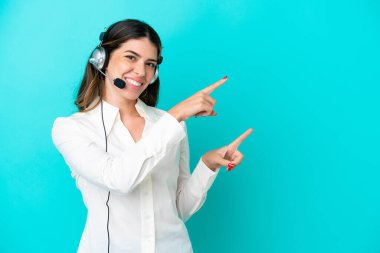 Telemarketer Italian woman working with a headset isolated on blue background pointing finger to the side and presenting a product