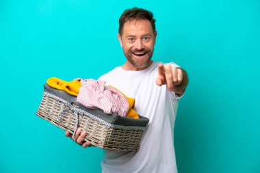 Middle age woman holding a clothes basket isolated on blue background surprised and pointing front