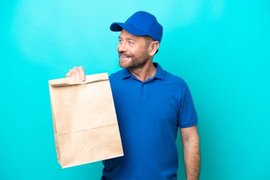 Middle age man taking a bag of takeaway food isolated on blue background looking to the side and smiling
