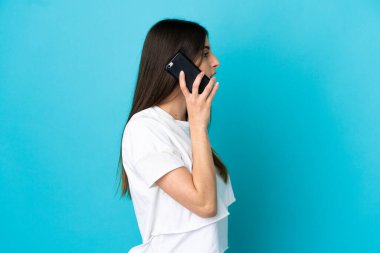 Mavi arka planda izole edilmiş genç beyaz bir kadın cep telefonuyla sohbet ediyor.