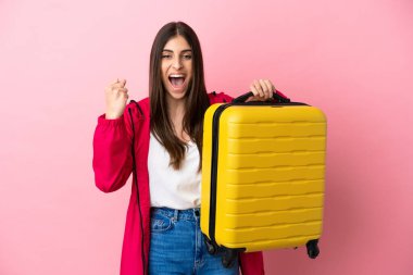 Pembe arka planda izole edilmiş genç beyaz kadın seyahat valiziyle geziyor.