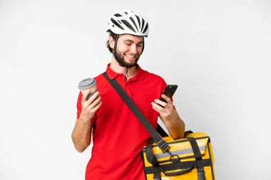 Termal sırt çantalı genç teslimatçı, izole edilmiş beyaz arka plan üzerinde kahve ve cep telefonu tutuyor.