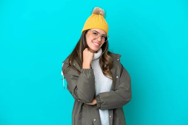 Young Italian woman wearing winter jacket and hat isolated on blue background with glasses and smiling