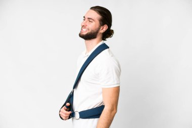 Young handsome man with broken arm and wearing a sling over isolated white background laughing in lateral position