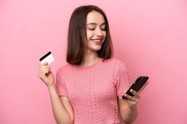Pembe arka planda izole edilmiş genç Ukraynalı bir kadın kredi kartıyla cep telefonuyla alışveriş yapıyor.