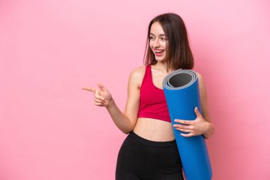 Young sport Ukrainian woman going to yoga classes while holding a mat isolated on pink background pointing finger to the side and presenting a product