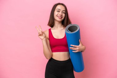 Young sport Ukrainian woman going to yoga classes while holding a mat isolated on pink background smiling and showing victory sign