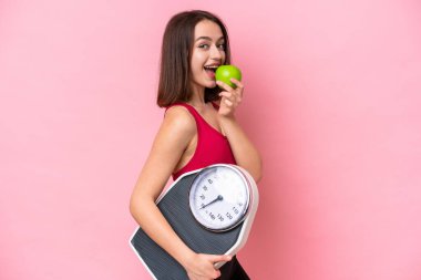Young Ukrainian woman isolated on pink background with weighing machine and with an apple