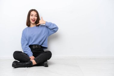 Young Ukrainian woman sitting on the floor isolated on white background making phone gesture. Call me back sign