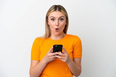 Beyaz arka planda izole edilmiş genç beyaz bir kadın kameraya bakarken şaşırmış bir ifadeyle cep telefonunu kullanıyor.