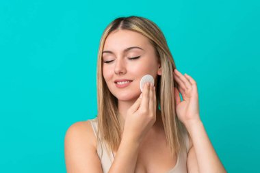 Yüzündeki makyajı silmek için mavi arka planda pamuklu pedi olan beyaz bir kadın.