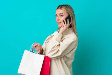 Kafkasyalı genç bir kadın mavi arka planda izole edilmiş. Elinde alışveriş poşetleri var ve cep telefonuyla arkadaşını arıyor.